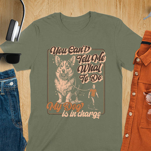 You Can't Tell Me What To Do My Dog Is In Charge text with man being walked on leash by corgi in vintage retro frame graphic on military green t-shirt