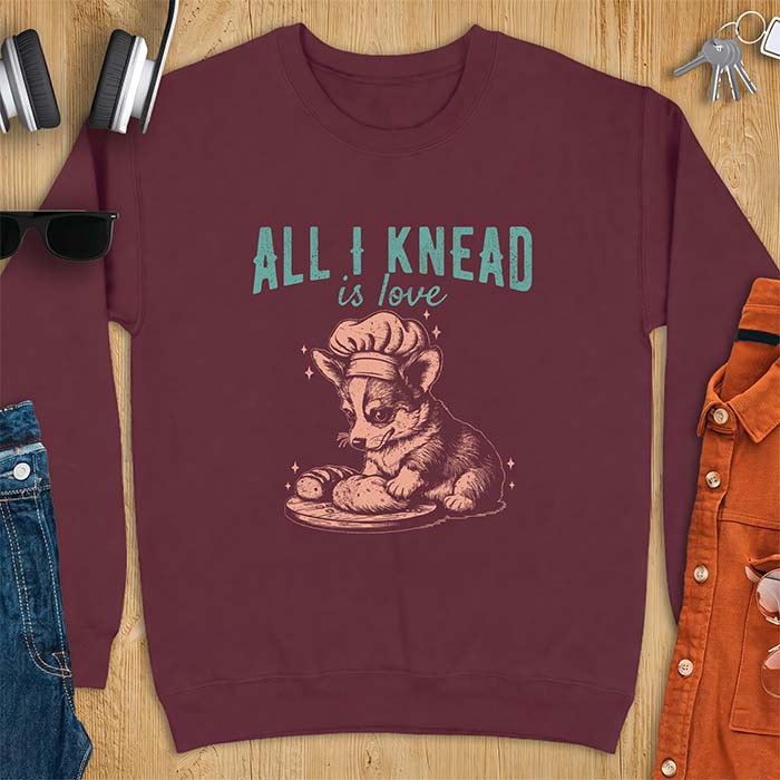 Maroon sweatshirt with a graphic of a corgi puppy wearing a chef's hat kneading a loaf of bread at a cutting board with sparkles around it, featuring the text All I Knead Is Love.