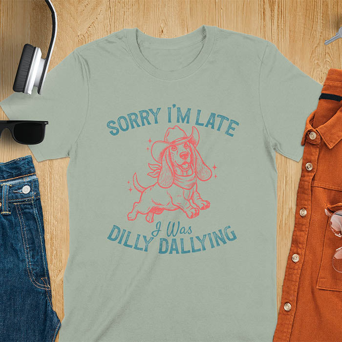 Sage green t-shirt featuring a galloping basset hound wearing a cowboy hat with long floppy ears flowing, text reads Sorry I'm Late I Was Dilly Dallying