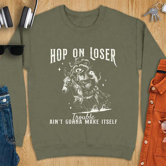 Military Green sweatshirt with a western graphic of a raccoon in a cowboy hat riding a donkey and an opossum riding along behind him under a scatter of stars and the text Hop On Loser, Trouble Ain't Gonna Make Itself