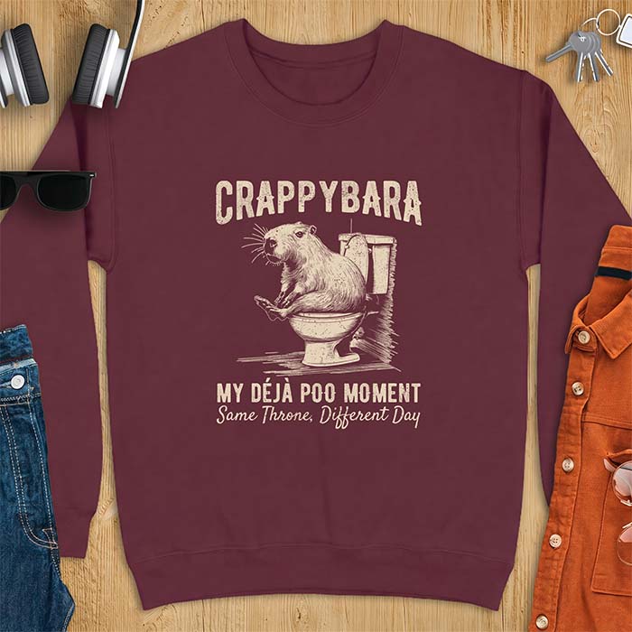 Maroon sweatshirt with a toilet humor graphic of a capybara sitting on an old-fashioned toilet, printed with the words Crappybara, My Deja Poo Moment, Same Throne, Different Day.