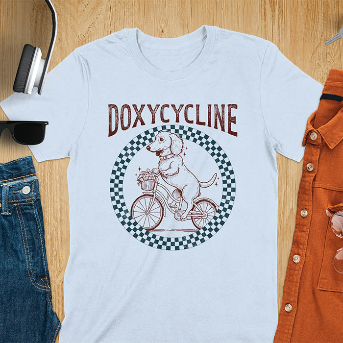 Light blue Dachshund t-shirt with 'DOXYCYCLINE' text and cute wiener dog riding bicycle in checkered circle design