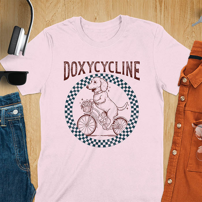 Light Pink Dachshund t-shirt with 'DOXYCYCLINE' text and cute wiener dog riding bicycle in checkered circle design