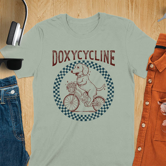 Sage green Dachshund t-shirt with 'DOXYCYCLINE' text and cute wiener dog riding bicycle in checkered circle design
