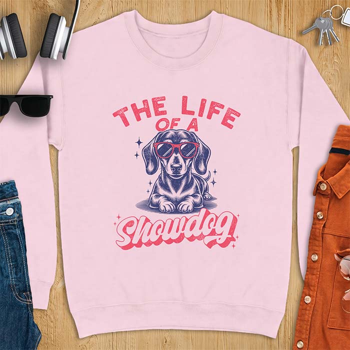 Light Pink sweatshirt with a graphic of a Dachshund wearing red sunglasses lying down surrounded by sparkles, with the text The Life Of A Showdog