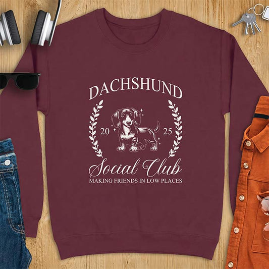 Maroon sweatshirt with a vintage crest graphic of a dachshund standing inside a laurel wreath alongside the text Dachshund, Social Club, 2025, Making Friends In Low Places.