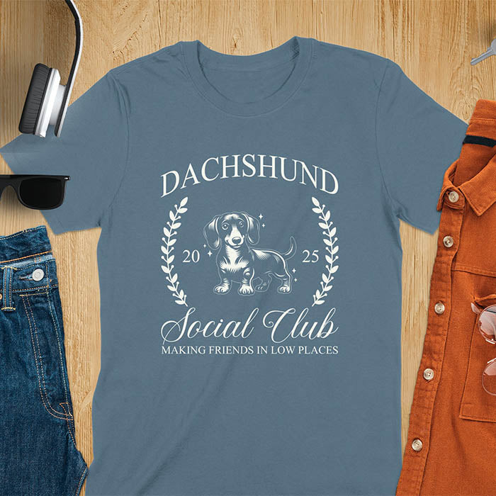 Indigo blue T-shirt with a playful dachshund graphic, laurel branches, and text reading "Dachshund Social Club," "EST 2024," and "Making Friends in Low Places." Vintage-inspired design perfect for dog lovers.