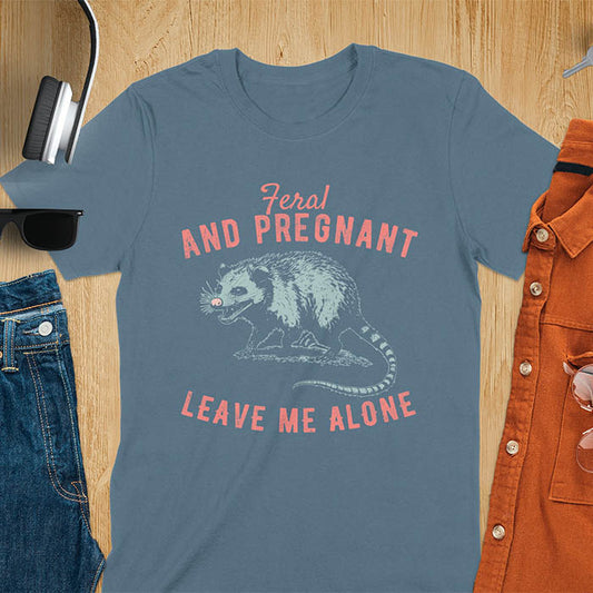 A indigo blue color t-shirt featuring An upset-looking opossum showing its fangs. Text: "Feral AND PREGNANT LEAVE ME ALONE". This funny maternity tee for pregnancy humor is ideal for expecting mom gift & mommy to be gift. Supports feral pregnancy, pregnancy announce, expecting baby, pregnancy keepsake. A unique new mama shirt, maternity apparel & gift mom hers.