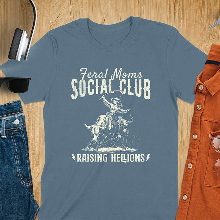 A indigo blue t-shirt with a cowgirl riding a bucking bull. Text: Feral Moms SOCIAL CLUB above and Raising HELLIONS below. This unhinged t-shirts design features a cowgirl mom on a rodeo bucking bull, a weird meme shirt for a feral mom gift. A retro inspired design, one of those shirts that go hard, a mama outfit or feral wife gift. A mom gift or gift for new mom for a western mama.