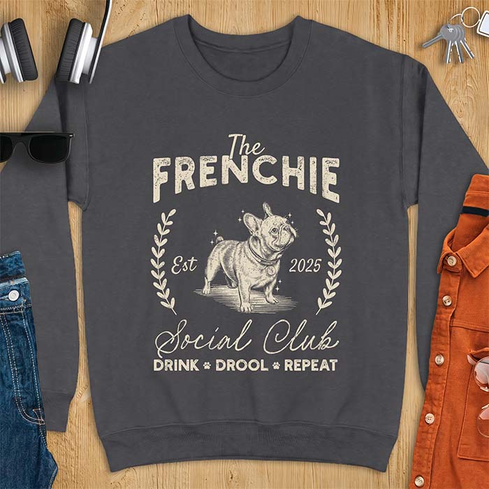 Dark Heather sweatshirt with a vintage badge graphic of a French Bulldog wearing a collar and tag framed by a laurel wreath, and the text The Frenchie, Est 2025, Social Club, Drink Drool Repeat