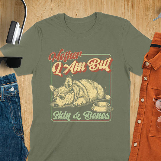 Chubby Corgi lying next to an empty bowl with a sulky expression, with the text Mother I Am But Skin and Bones in retro typography on a military green t-shirt