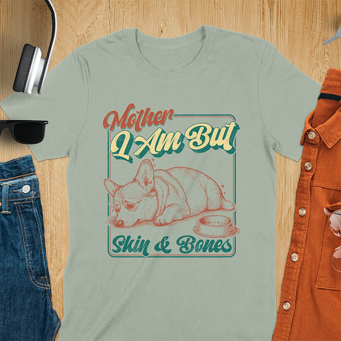 Chubby Corgi lying next to an empty bowl with a sulky expression, with the text Mother I Am But Skin and Bones in retro typography on a sage green t-shirt
