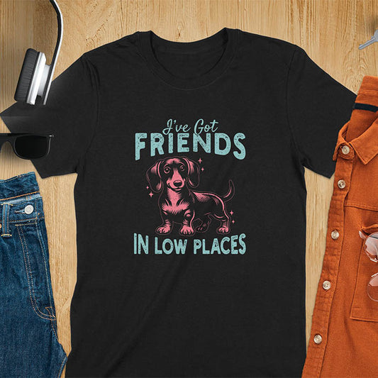 This T-shirt features a vintage-style line art of a happy dachshund looking straight at you with its tail held high. The top text reads, I've got friends, and below it says, in low places. Overall in a retro color palette with a distressed texture, on a black tee.