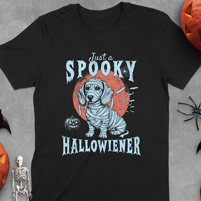 Black t-shirt featuring a cute dachshund puppy wrapped in mummy bandages with text 'Just a Spooky Hallowiener', jack-o'-lantern pumpkin, red moon and flying bats in background