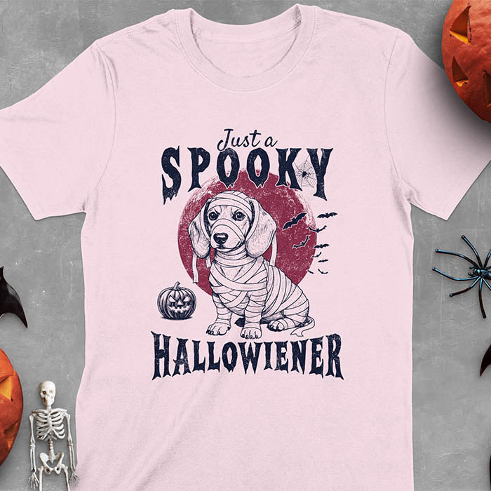 Light Pink t-shirt featuring a cute dachshund puppy wrapped in mummy bandages with text 'Just a Spooky Hallowiener', jack-o'-lantern pumpkin, red moon and flying bats in background