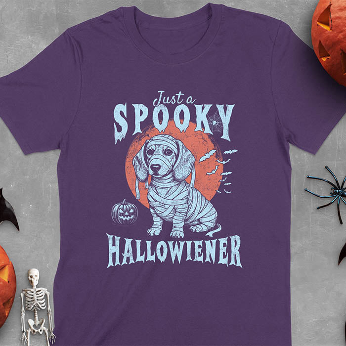 Purple t-shirt featuring a cute dachshund puppy wrapped in mummy bandages with text 'Just a Spooky Hallowiener', jack-o'-lantern pumpkin, red moon and flying bats in background