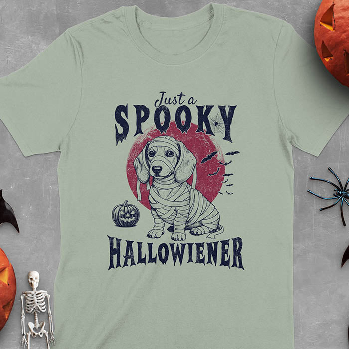 Sage green t-shirt featuring a cute dachshund puppy wrapped in mummy bandages with text 'Just a Spooky Hallowiener', jack-o'-lantern pumpkin, red moon and flying bats in background