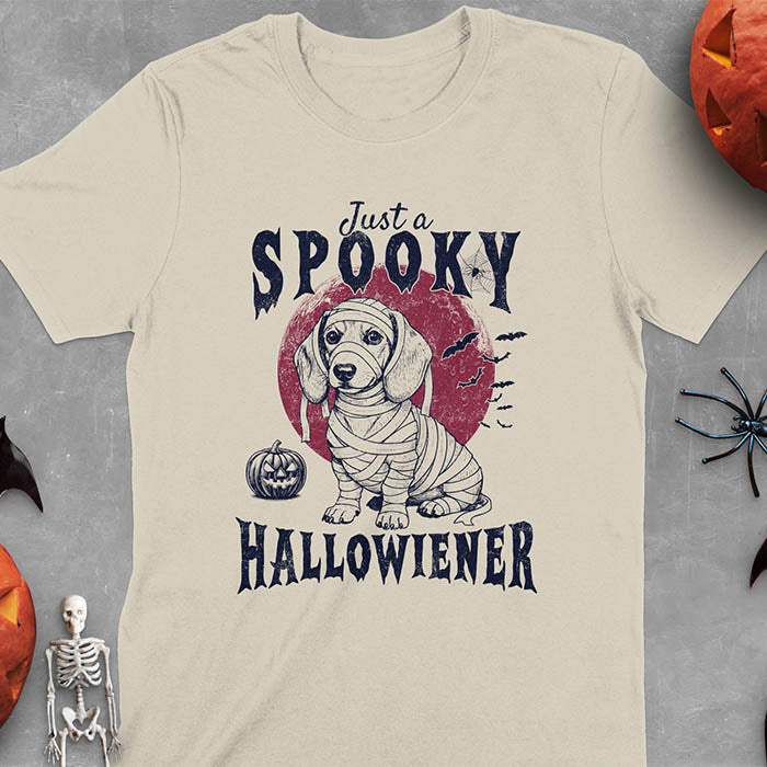 Sand color t-shirt featuring a cute dachshund puppy wrapped in mummy bandages with text 'Just a Spooky Hallowiener', jack-o'-lantern pumpkin, red moon and flying bats in background