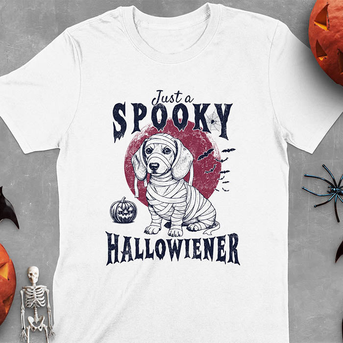 White t-shirt featuring a cute dachshund puppy wrapped in mummy bandages with text 'Just a Spooky Hallowiener', jack-o'-lantern pumpkin, red moon and flying bats in background