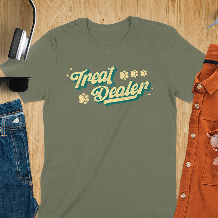 Military Green t-shirt with 'Treat Dealer' text on a wooden surface with sunglasses, jeans, and an orange jacket.