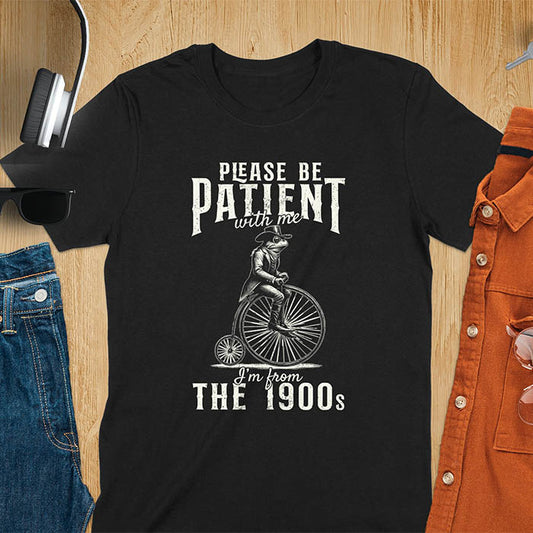 A black t-shirt with a frog riding a penny-farthing bicycle. Text: "PLEASE BE PATIENT with me" above and "I'm from THE 1900s" below. This The 1900s shirt has a vintage graphic tee and retro aesthetic. A genz Meme shirt and weird silly shirt, a funny gift friend. A gift for husband, funny grandpa gift, or dad tee-shirt. An animal person gift, a 90s country shirt, a mom gift option.