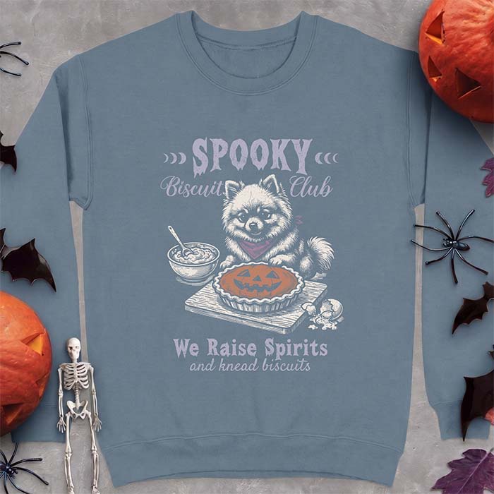 Indigo Blue sweatshirt with a Halloween graphic of a Pomeranian wearing a plaid bandana sitting behind a jack-o'-lantern pumpkin pie on a cutting board with a mixing bowl, spoon and a cracked egg nearby and the text Spooky Biscuit Club, We Raise Spirits and knead biscuits