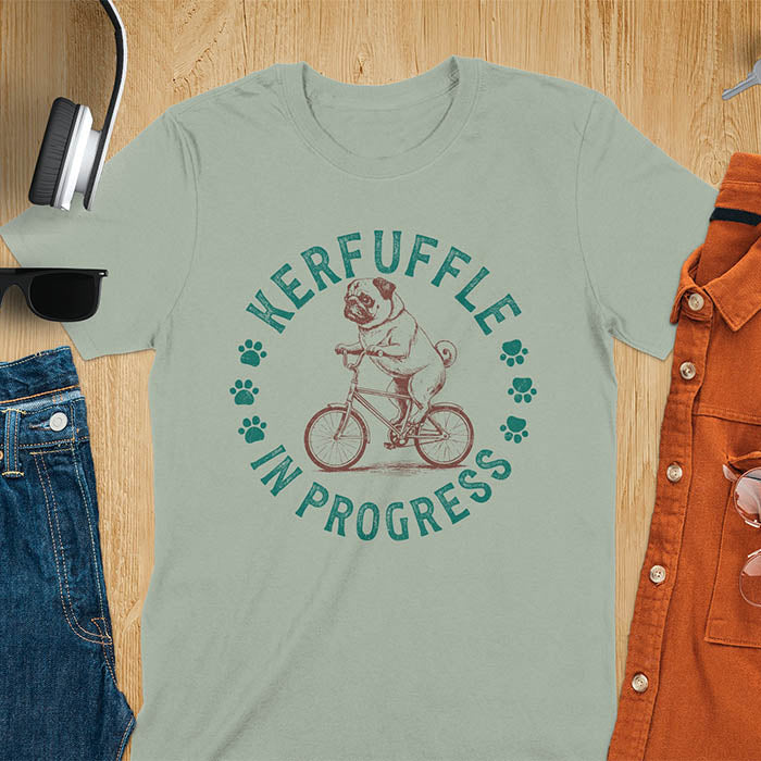 Pug on bicycle sketch-style graphic with Kerfuffle In Progress text and paw prints on sage green t-shirt