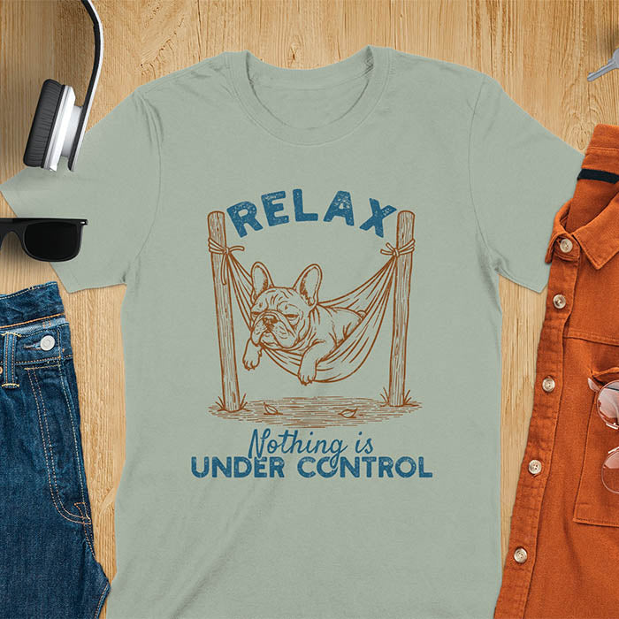 Sage green t-shirt with vintage-inspired illustration. Retro graphic features French bulldog relaxing in hammock with 'Relax' and 'Nothing is Under Control' text in distressed typography. Quirky zen humor meme design.