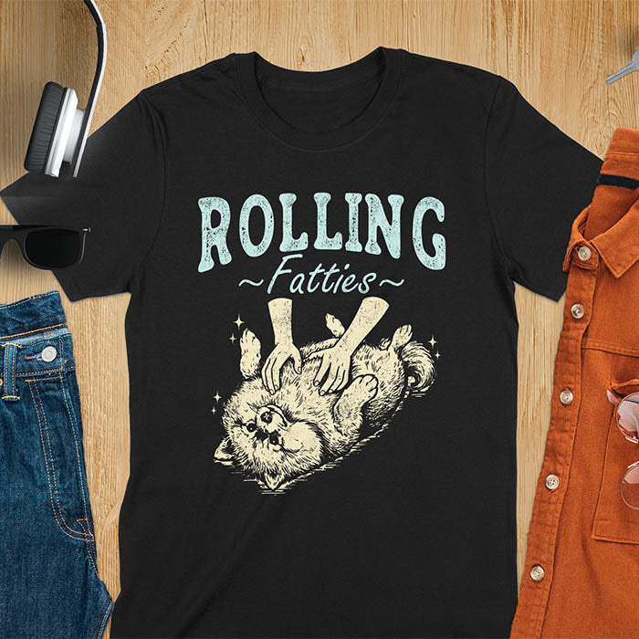 Black t-shirt with vintage-inspired illustration. Retro graphic features fluffy Pomeranian rolling on back with 'Rolling Fatties' text in distressed typography. Quirky chubby dog meme design.