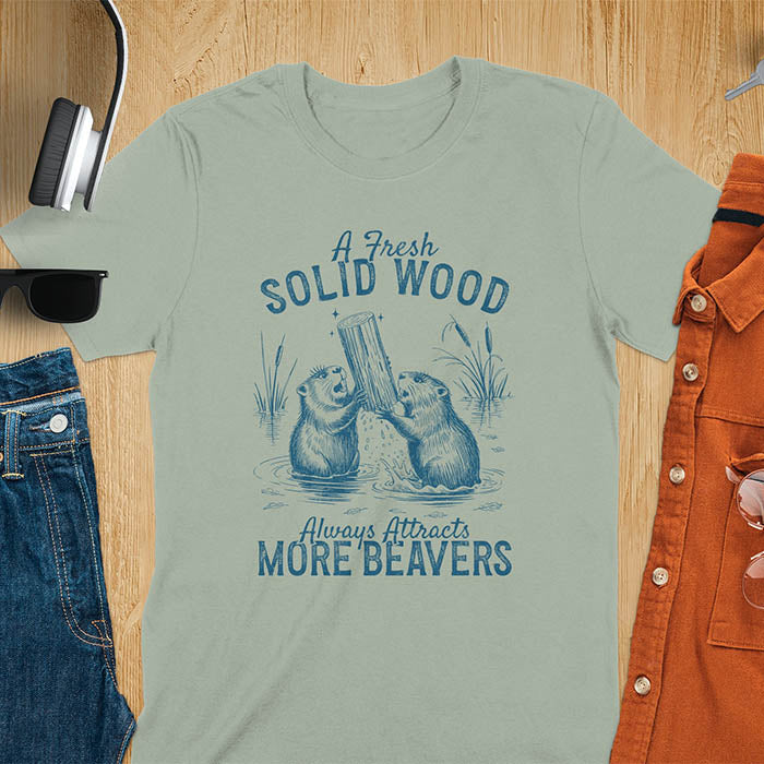 Sage green t-shirt with vintage-style illustration of two beavers in water fighting over a wooden log, featuring text 'A Fresh Solid Wood Always Attracts More Beavers' in rustic lettering