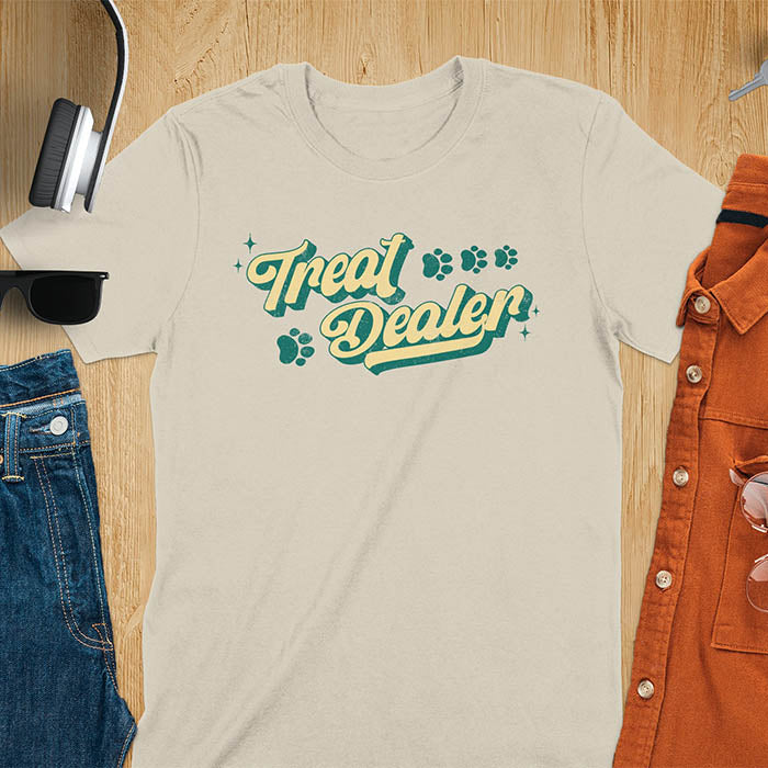 Sand color t-shirt with Treat Dealer text in retro script typography with green and yellow colors and paw print decorations