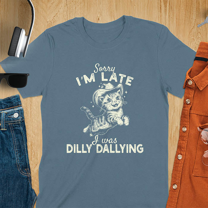 A indigo blue t-shirt with a tabby cat wearing a cowboy hat. Text: Sorry I'M LATE above and I was DILLY DALLYING below. This cat lover shirt features cat cuteness gift and kawaiiness cuteness. A sarcastic meme shirt about dilly dallying, a funny genz meme with funny animals. This cowboy cat design has retro aesthetics, a snarky humorous tee, an animal person gift or mom gift.