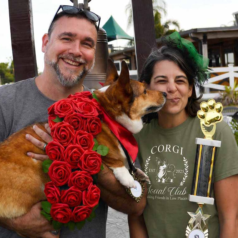 Tank the corgi with his owners at the Tampa Bay Downs Corgi Derby, the woman in a Corgi Social Club tee, holding a championship trophy