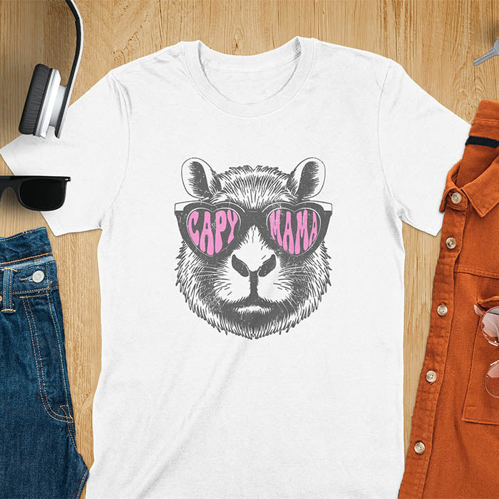 A t-shirt featuring a capybara face with pink heart-shaped sunglasses that spell CAPY MAMA. Below the capybara, Meagan is written in a pink, script font. This vintage mama shirt design is a retro mom tee-shirt perfect for mama mom life. A trendy mama tee for an expecting mom or pregnant mom gift, ideal for a pregnancy announce or pregnancy reveal. A great mom gift hers or mother days gift, or a new mom gift for a gift for wife. Personalization is available by changing the Meagan name.