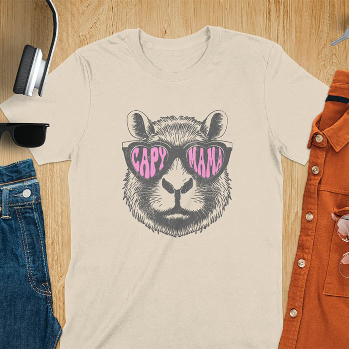 A t-shirt featuring a capybara face with pink heart-shaped sunglasses that spell CAPY MAMA. Below the capybara, Meagan is written in a pink, script font. This vintage mama shirt design is a retro mom tee-shirt perfect for mama mom life. A trendy mama tee for an expecting mom or pregnant mom gift, ideal for a pregnancy announce or pregnancy reveal. A great mom gift hers or mother days gift, or a new mom gift for a gift for wife. Personalization is available by changing the Meagan name.