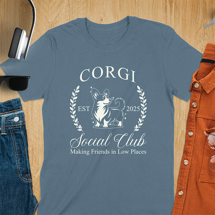 Our charming Corgi Social Club t-shirt features a delightful illustration of a corgi surrounded by laurel branches, with playful text that reads CORGI Social Club and Making friends in low places.