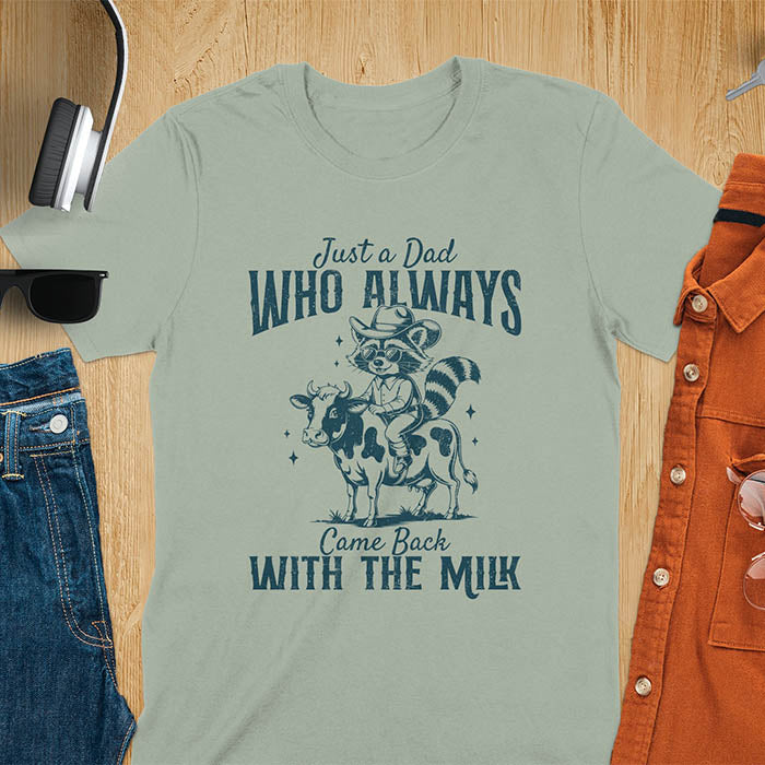 A t-shirt with a raccoon wearing a cowboy hat and sunglasses, riding a cow. Text: Just a Dad WHO ALWAYS above and Came Back WITH THE MILK below. This funny raccoon shirt features funny graphics, a dad t shirt for a funny fathers day or fathers day gift. A funny shirt for dad or gift for dad, a husband gift or father in law gifts. Could be used for a dad announcement, expectant dad, or godfather proposal.