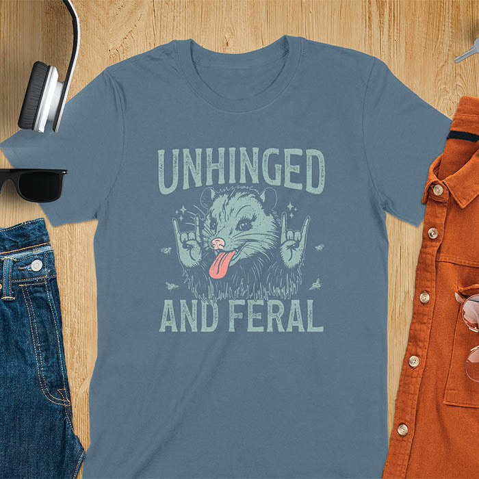 A t-shirt featuring a opossum with its tongue sticking out, throwing rock-on hand gestures. Small flies surround the opossum. Text: UNHINGED above and AND FERAL below in a distressed font. This unhinged shirt features a funny opossum design, a weird meme shirt or weird t shirt for unhinged gifts. A sarcasm shirt with a sassy shirt attitude, a memeing shirt or genz meme for an animal person gift.