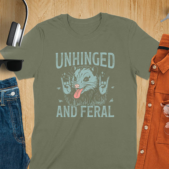 A t-shirt featuring a opossum with its tongue sticking out, throwing rock-on hand gestures. Small flies surround the opossum. Text: UNHINGED above and AND FERAL below in a distressed font. This unhinged shirt features a funny opossum design, a weird meme shirt or weird t shirt for unhinged gifts. A sarcasm shirt with a sassy shirt attitude, a memeing shirt or genz meme for an animal person gift.