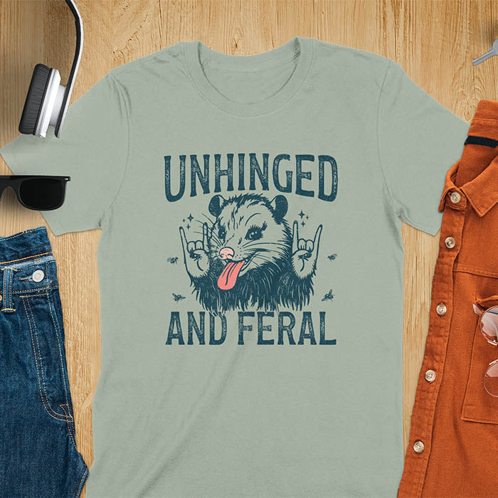 A t-shirt featuring a opossum with its tongue sticking out, throwing rock-on hand gestures. Small flies surround the opossum. Text: UNHINGED above and AND FERAL below in a distressed font. This unhinged shirt features a funny opossum design, a weird meme shirt or weird t shirt for unhinged gifts. A sarcasm shirt with a sassy shirt attitude, a memeing shirt or genz meme for an animal person gift.