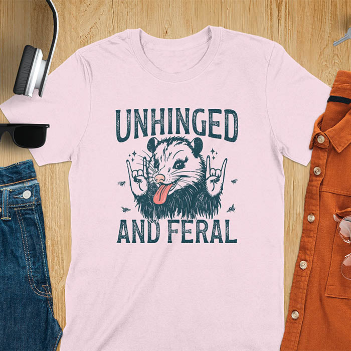 A t-shirt featuring a opossum with its tongue sticking out, throwing rock-on hand gestures. Small flies surround the opossum. Text: UNHINGED above and AND FERAL below in a distressed font. This unhinged shirt features a funny opossum design, a weird meme shirt or weird t shirt for unhinged gifts. A sarcasm shirt with a sassy shirt attitude, a memeing shirt or genz meme for an animal person gift.