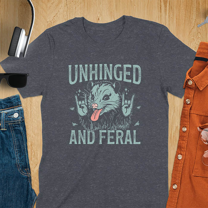 A t-shirt featuring a opossum with its tongue sticking out, throwing rock-on hand gestures. Small flies surround the opossum. Text: UNHINGED above and AND FERAL below in a distressed font. This unhinged shirt features a funny opossum design, a weird meme shirt or weird t shirt for unhinged gifts. A sarcasm shirt with a sassy shirt attitude, a memeing shirt or genz meme for an animal person gift.