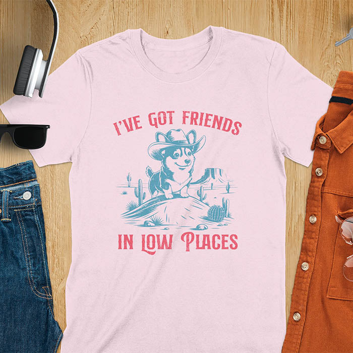 A t-shirt with a corgi wearing a cowboy hat sitting in a desert scene with cacti. Text: "I've Got FRIENDS" above and "IN LOW PLACES" below. This retro dog shirt is a corgi shirt and funny dog shirt, a vintage dog tee. A cowboy tshirt for a dog dad shirt and pet owner gift. An animal person gift with cuteness puppy, a meme graphic tee, a mom gift option.