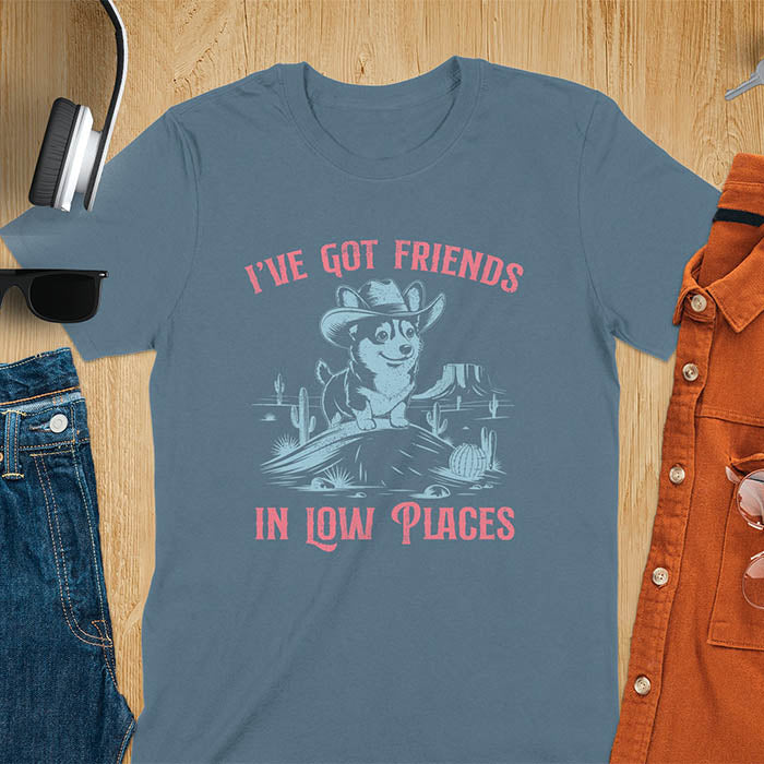 A t-shirt with a corgi wearing a cowboy hat sitting in a desert scene with cacti. Text: "I've Got FRIENDS" above and "IN LOW PLACES" below. This retro dog shirt is a corgi shirt and funny dog shirt, a vintage dog tee. A cowboy tshirt for a dog dad shirt and pet owner gift. An animal person gift with cuteness puppy, a meme graphic tee, a mom gift option.