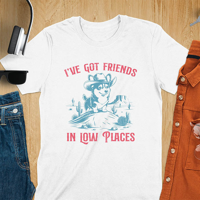 A t-shirt with a corgi wearing a cowboy hat sitting in a desert scene with cacti. Text: "I've Got FRIENDS" above and "IN LOW PLACES" below. This retro dog shirt is a corgi shirt and funny dog shirt, a vintage dog tee. A cowboy tshirt for a dog dad shirt and pet owner gift. An animal person gift with cuteness puppy, a meme graphic tee, a mom gift option.