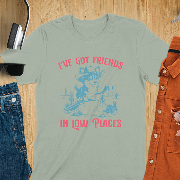 A t-shirt with a corgi wearing a cowboy hat sitting in a desert scene with cacti. Text: "I've Got FRIENDS" above and "IN LOW PLACES" below. This retro dog shirt is a corgi shirt and funny dog shirt, a vintage dog tee. A cowboy tshirt for a dog dad shirt and pet owner gift. An animal person gift with cuteness puppy, a meme graphic tee, a mom gift option.