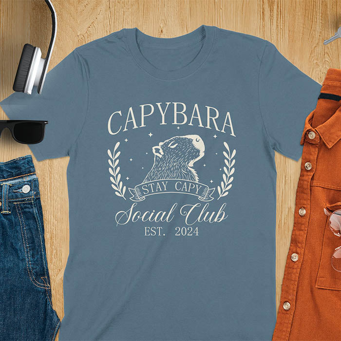 Blue t-shirt with 'Capybara Stay Capy Social Club' design on a wooden surface with accessories.