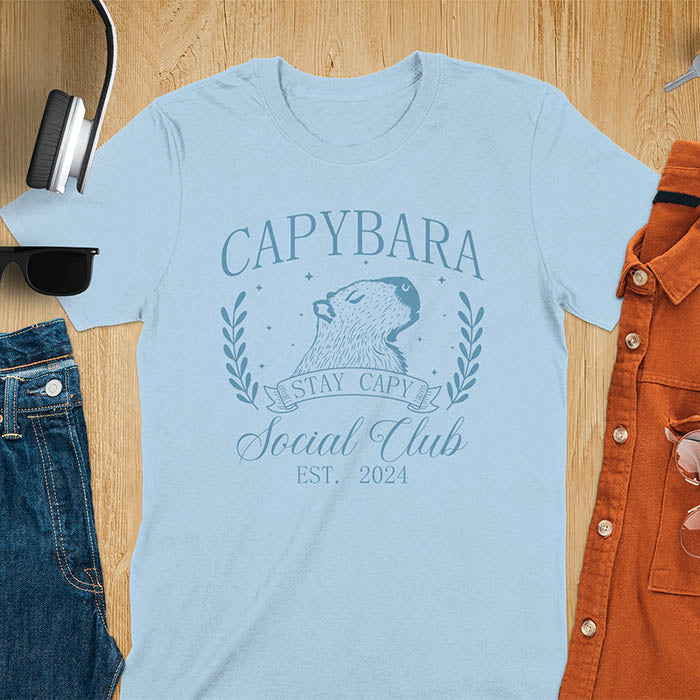 Light blue t-shirt with capybara design and text on a wooden surface with accessories.  Stay capy slogan.