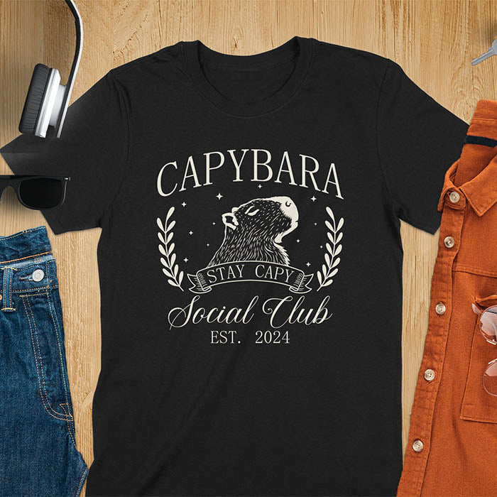 Black t-shirt with 'Capybara Stay Capy Social Club' design on a wooden surface with accessories. 