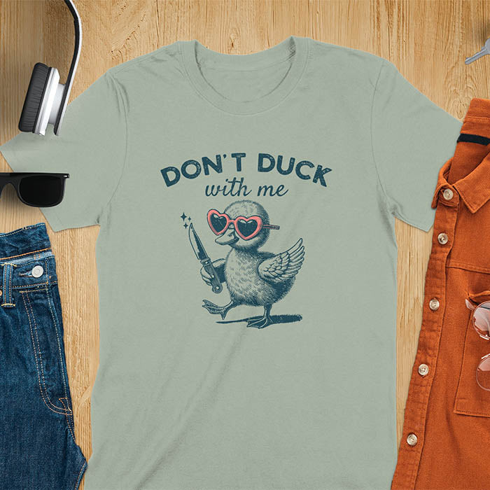 A T-shirt with a cartoon duck wearing heart-shaped sunglasses and holding a knife. The text above reads DON'T DUCK with me. The design is humorous and edgy, combining playful imagery with a bold statement.
